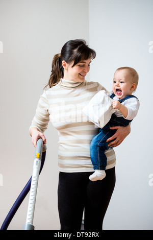 Reinigung des Zimmers - Mutter mit Staubsauger hält ihr Baby auf Händen Stockfoto