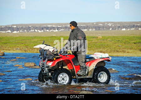 Mann von den Inuit, die Fahrrad fahren Quad, ATV, durch einen Fluss in der Tundra auf Victoria Island, früher Holman Island Stockfoto