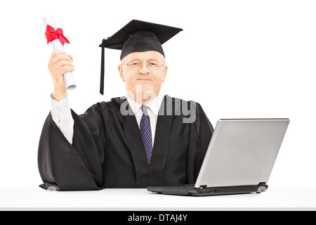 Reifer Mann in Graduierung Kleid posiert mit Diplom und Laptop auf Tisch Stockfoto