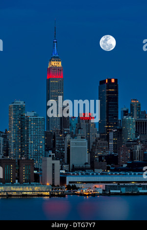 Der Vollmond steigt über die Skyline von New York City (NYC) während die Dämmerstunde. Ein Blick aus New Jersey über den Hudson River mit dem Empire State Building in rot, weiß und blau beleuchtet. Stockfoto