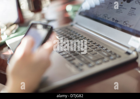 Eine Frau sitzend am Schreibtisch hält ein Smartphone mit einem Laptop-Computer New Hope, Pennsylvania USA Stockfoto