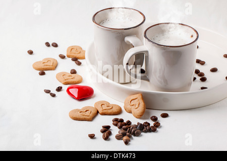 Zwei Tassen Cappuccino serviert mit Gewürzen und Cookies als Herz und ein rotes Herz auf weißen Textilien. Stockfoto