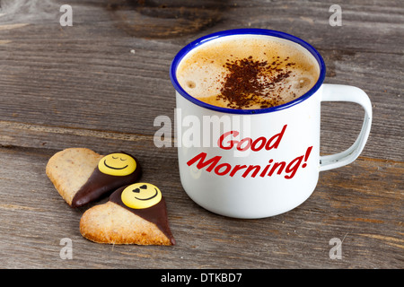 Emaille Becher mit heißem Kaffee und zwei Kekse in Herzform auf einem rustikalen Holzbrett mit dem Wort 'Guten Morgen' auf der Tasse Stockfoto