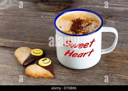 Emaille Becher mit heißem Kaffee und zwei Kekse in Herzform auf einem rustikalen Holzbrett mit dem Wort "Sweet Heart" auf der Tasse Stockfoto