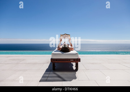 Frau liest auf Liegestuhl am Pool mit Blick auf Meer Stockfoto