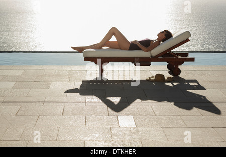 Frau, die ein Sonnenbad im Liegestuhl am Pool mit Blick auf Meer Stockfoto