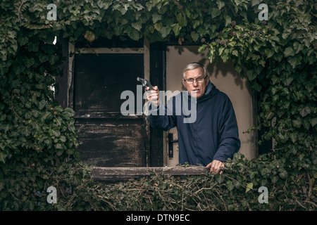 Wütend senior Mann mit einer Waffe zum Schutz seines Eigentums, steht man vor alten verwilderten Kabine gestikulieren. Stockfoto