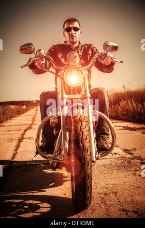 Biker-Mann trägt eine Lederjacke und eine Sonnenbrille sitzt auf seinem Motorrad, Blick auf den Sonnenuntergang. Stockfoto