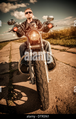 Biker-Mann trägt eine Lederjacke und eine Sonnenbrille sitzt auf seinem Motorrad, Blick auf den Sonnenuntergang. Stockfoto