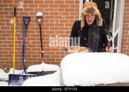 Ein Mann leitet durch die Hintertür mit einem Tablett mit Huhn und starrt ungläubig auf seinen Grill begraben im Schnee. Stockfoto