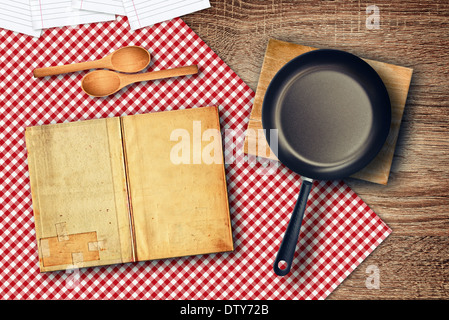 Zubereitung von Speisen am Küchentisch. Diverse Küchenutensilien auf Tischplatte - Löffel, Rezeptbuch und Pfanne. Stockfoto