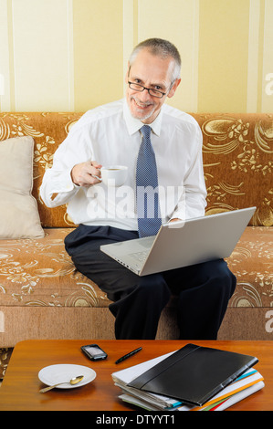 Ein hübscher senior Geschäftsmann arbeiten mit Computer zu Hause bereit, eine heiße Tasse schwarzen Kaffee zu trinken Stockfoto