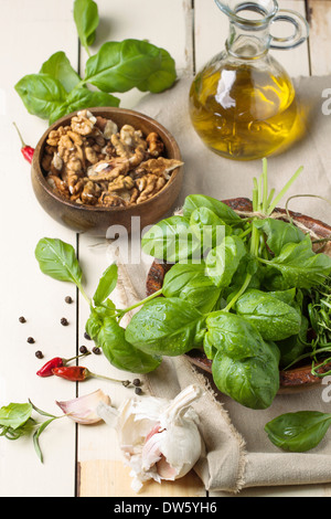 Jede Menge frisches Basilikum, Schale mit Nüssen, Pfeffer, Knoblauch und Glasflasche Olivenöl auf weißer Holztisch serviert. Serien ansehen Stockfoto