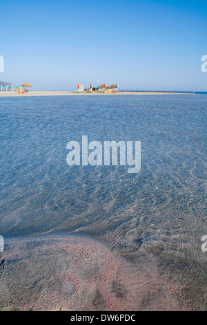 Sand mit roten Fragmente von Seeigel, Muscheln, klaren blauen Wasser und entfernte Sonne Schirme auf Elafonissos Strand auf Kreta Stockfoto