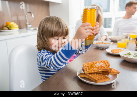 Fröhlicher Junge Gießen Honig auf Waffeln beim Frühstück mit der Familie Stockfoto