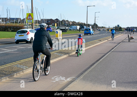 Vater und Sohn, sonnige Radfahrer im Winter, auf einer speziellen Fahrradspur am Straßenrand neben dem Verkehr am Meer in Southend auf der Sea Essex England UK Stockfoto
