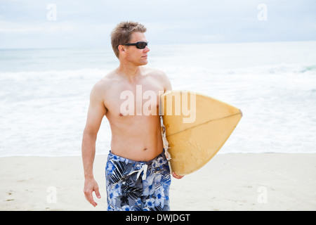 Entspannt und konzentriert aussehende männliche Surfer zu Fuß am Strand während des Tragens Surfbrett Stockfoto