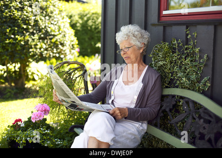 Entspannte alte Frau sitzt auf der Bank im Garten lesen Zeitung Stockfoto