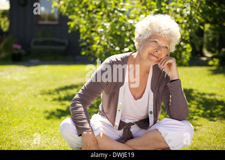 Ältere Frau sitzen im Freien auf dem Rasen wegschauen und denken. Ältere Frau Entspannung in Hinterhof Garten Tagträumen Stockfoto