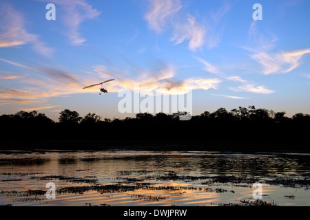 Ein Microlight Flugzeug fliegt über einen Kanal in das Okavango Delta in Botswana bei Sonnenuntergang Stockfoto