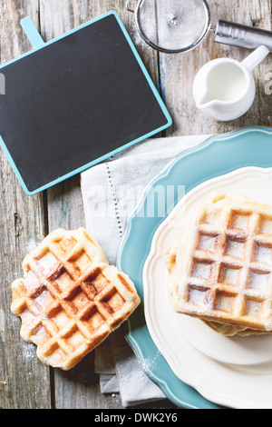 Draufsicht auf Teller mit frischen Waffeln serviert mit leeren schwarzen Tafel über alten Holztisch. Stockfoto