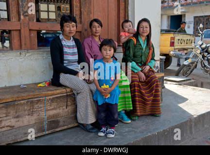 Mütter in Trachten mit Kindern sitzen auf einer Bank in der Hauptstraße von Paro, Bhutan Stockfoto