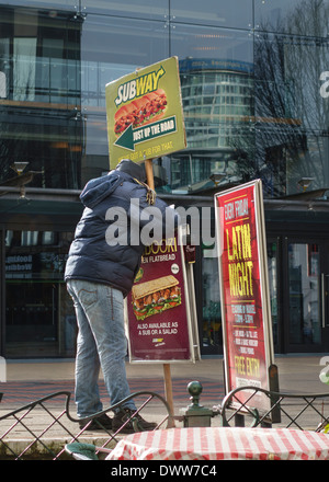 Ein Mann hält ein Brett, das die Subway Fast-Food-Kette im Stadtzentrum von Birmingham, Großbritannien, wirbt. Das Rotunda-Gebäude spiegelt sich im Hintergrund wider Stockfoto