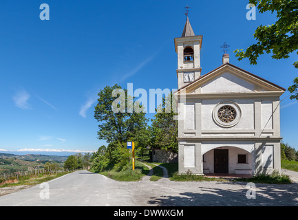 Weiße Kapelle am Straßenrand und einspurige Straße unter blauem Himmel im Piemont, Norditalien. Stockfoto