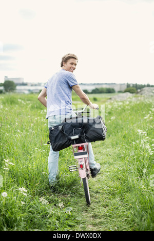 Hintere Ansicht Porträt junger Mann mit Tasche und Fahrrad stehen auf Feld Stockfoto