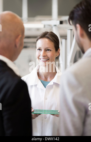 Lächelnd Ingenieur zeigt Maschinenteil für Geschäftsleute im Werksgelände Stockfoto