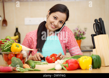Lächelnde junge Frau Schneiden von Gemüse und Gespräch am Handy Stockfoto