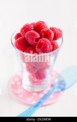 Nahaufnahme von frischen roten Himbeeren im Glas mit blauen Plastiklöffel ruht auf kleinen rosa Teller auf der Leuchtfläche Stockfoto