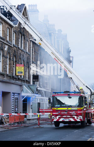 Feuerwehrmann in gelben Helm steht auf rot Motor an der Basis der Teleskopleiter von rauchig, Feuer beschädigte Gebäude - Stadt Harrogate, England, Großbritannien Stockfoto