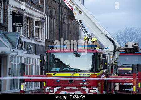 2 Feuerwehrmänner in gelb Helme, stehend auf rot Motor oder Fahrzeug durch Base der Teleskopleiter außerhalb Feuer beschädigte Gebäude - Harrogate, England, UK. Stockfoto