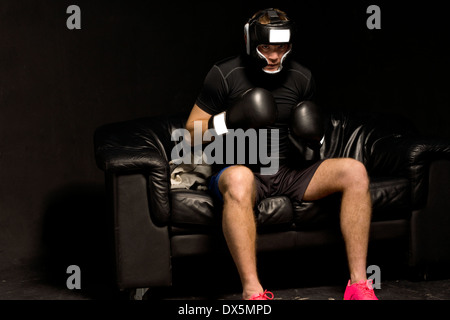 Boxer sitzt auf einer Couch, immer bereit für einen Kampf, Helm und Handschuhe anziehen, wie er in der Dunkelheit sitzt Stockfoto