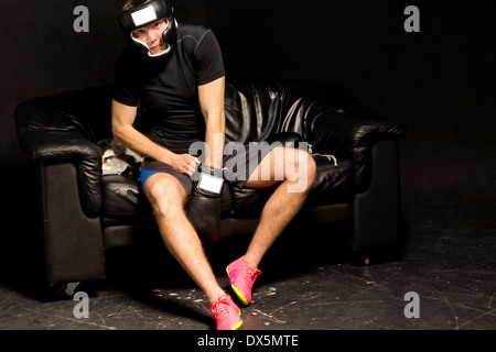 Junge Boxer passt seine Boxhandschuhe, als er sich für einen Kampf auf einer schwarzen Ledercouch auf einem dunklen Hintergrund sitzen bereitet Stockfoto