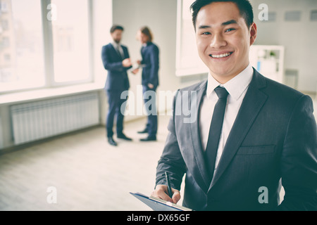 Close-up Portrait von ein junger erfolgreicher Manager mit Zwischenablage posiert auf Kamera, während seine Kollegen reden auf dem Hintergrund Stockfoto