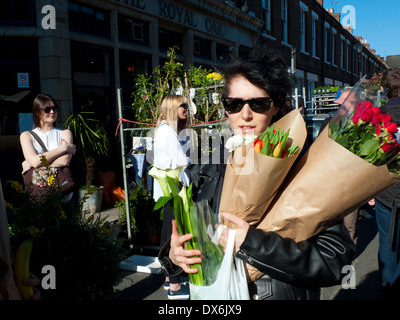 Frau, die Pflanze kaufen pflanzen Rosen Tulpen rote Rosen zu Fuß entlang der Straße in der Sonne an der Columbia Road Blumenmarkt London E2 UK KATHY DEWITT Stockfoto