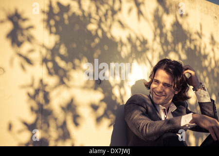 Modisch junger Mann sitzt mit einer Mauer im Schatten an einem sonnigen Tag Stockfoto