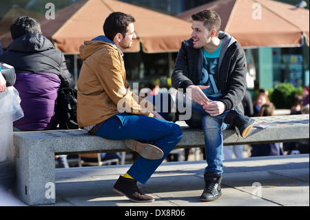 Zwei junge Männer sitzen auf einer Bank Konzert gekleidet in Winterkleidung sprechen, London, England, Vereinigtes Königreich. Stockfoto