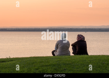 Freundinnen auf dem Rasen. Zwei Frauen, die Kopftücher tragen chat bei Sonnenuntergang am Ufer des Ottawa River. Stockfoto