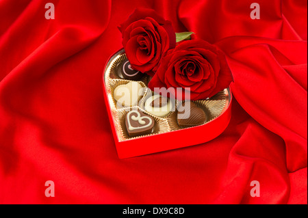 rote Rosen und köstliche Pralinen in goldenes Herz-Form-Geschenk-Box auf satin Hintergrund Stockfoto