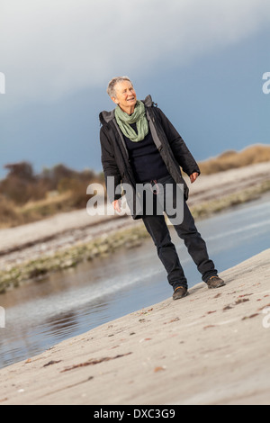 Beautiful senior Woman herumtollen am Strand schreitende zusammen mit ausgebreiteten Armen und einem Lächeln der Wertschätzung, wie sie Natur und die Freiheit von ihrem Ruhestand genießt Stockfoto