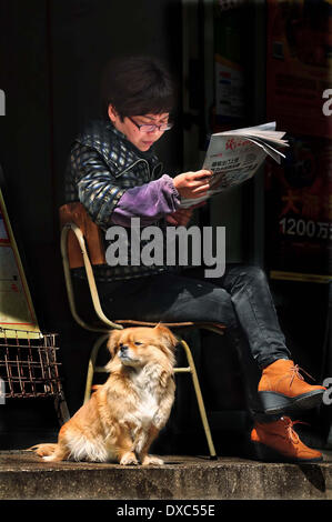 Ningbo, China Zhejiang Provinz. 23. März 2014. Ein Hund genießt Sonnenschein neben einer Frau, die Zeitung auf einer Straße in Ningbo, einer Hafenstadt im Osten Chinas Zhejiang Provinz, 23. März 2014 liest. © Hu Xuejun/Xinhua/Alamy Live-Nachrichten Stockfoto