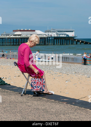 Frau sitzt auf einem kleinen Stuhl am Strand mit Cromer Pier im Hintergrund England Stockfoto