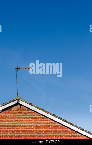 TV-Antenne / Antenne an Spitze der abfallenden Dach über roten Backsteinmauer gegen überwiegend blauen Himmel mit verschwommenen Wolken gefliest. Stockfoto