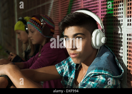 Close-up Portrait von Junge sitzt neben Wand, tragen von Kopfhörern und Musik hören, andere Kinder im Hintergrund, Deutschland Stockfoto