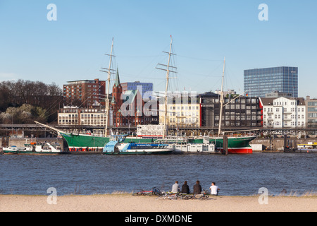 Gruppe von jungen Leuten sitzt am Strand gegenüber dem Museumsschiff Rickmer Rickmers in Hamburg, Deutschland. Stockfoto