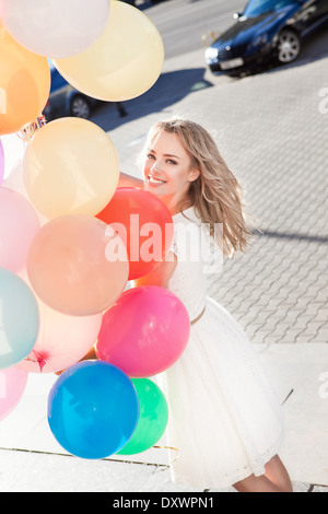 ziemlich lächelnde junge Frau im weißen Sommerkleid mit einer Reihe von bunten Luftballons an sonnigen Tag in der Stadt Stockfoto