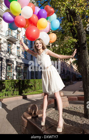 hübsche Frau mit langen Beinen in weisses Sommerkleid hält eine Reihe von bunten Luftballons steht auf Bank im Stadtpark Stockfoto